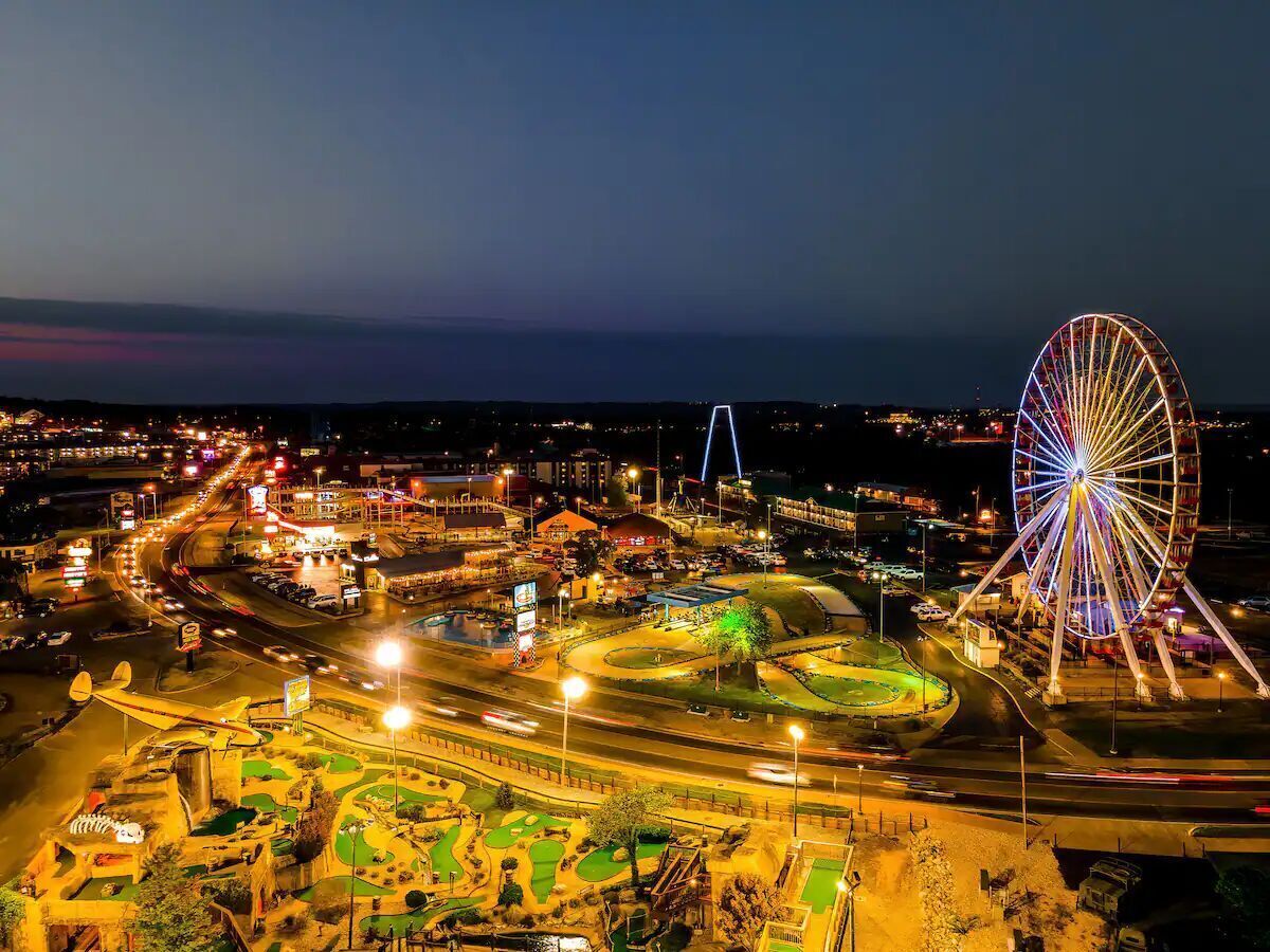 A large Ferris wheel with colorful lights, surrounded by a well-lit amusement park area with various rides and attractions.