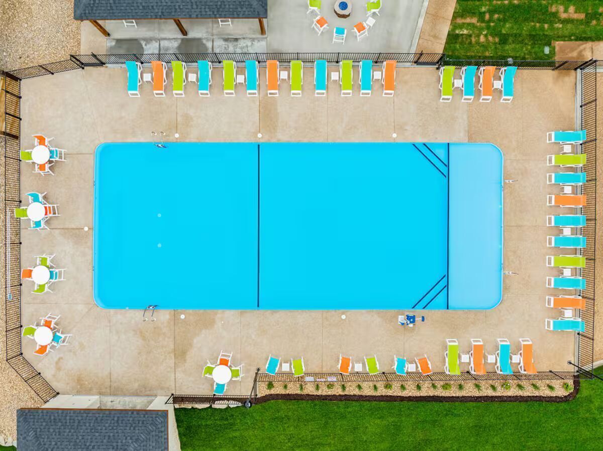 An aerial view of a rectangular swimming pool with adjacent lounge areas, surrounded by a fence and a grassy area.