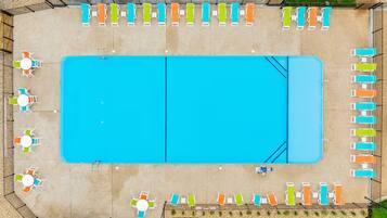 An aerial view of a rectangular swimming pool with adjacent lounge areas, surrounded by a fence and a grassy area.