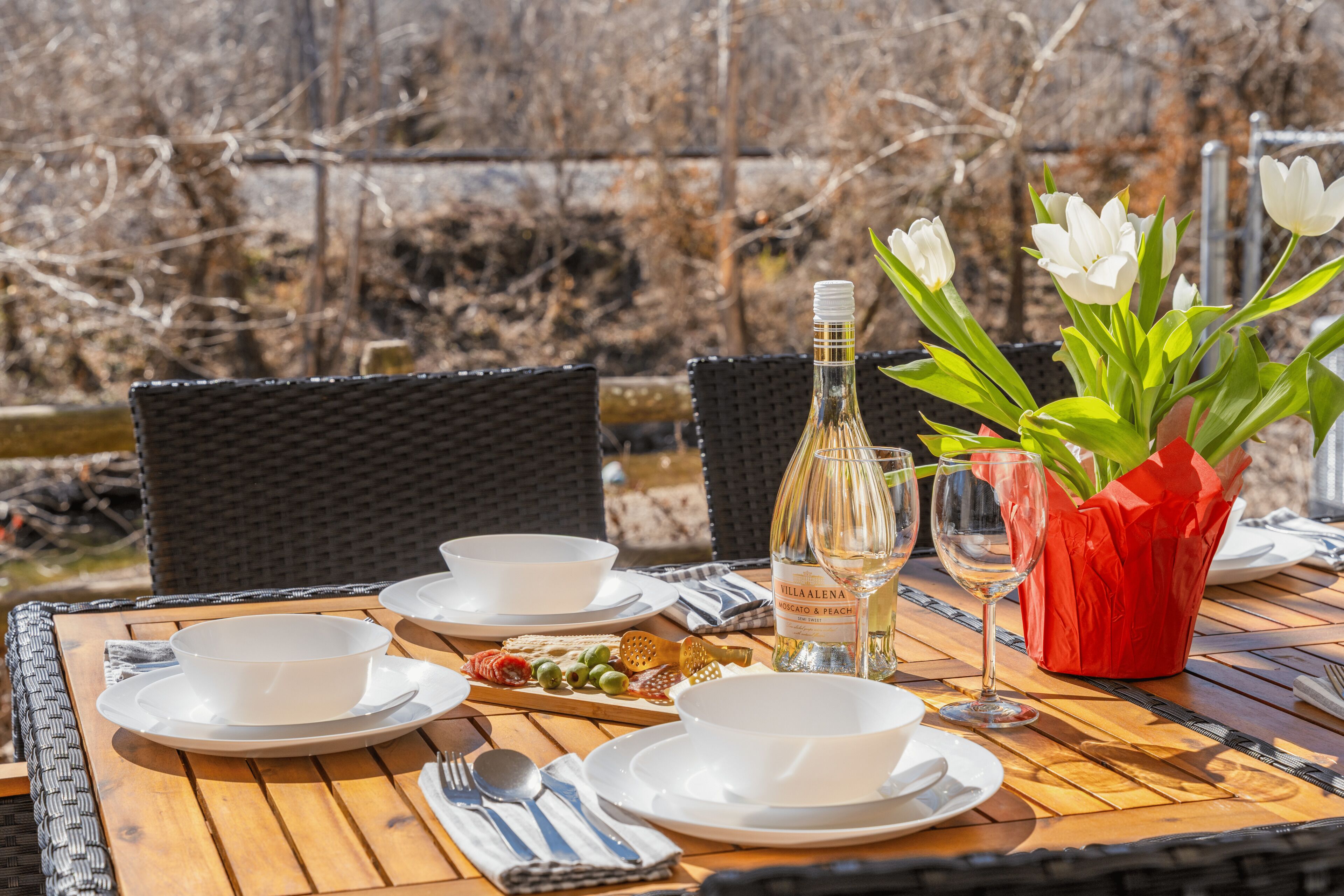 An outdoor dining table with plates, bowls, cutlery, a bottle of wine, and a vase with white flowers.
