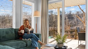 Una persona sentada en un sofá verde leyendo un libro, con una lámpara y una planta en maceta sobre una mesa frente a un ventanal corredizo que da a un área exterior con terraza de madera y sillas.