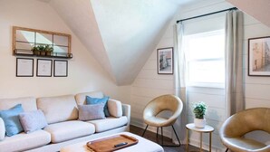 Une chambre à coucher confortable, aménagée en mezzanine, avec un plafond en pente, une grande fenêtre avec des rideaux, un canapé, deux fauteuils et une petite table.