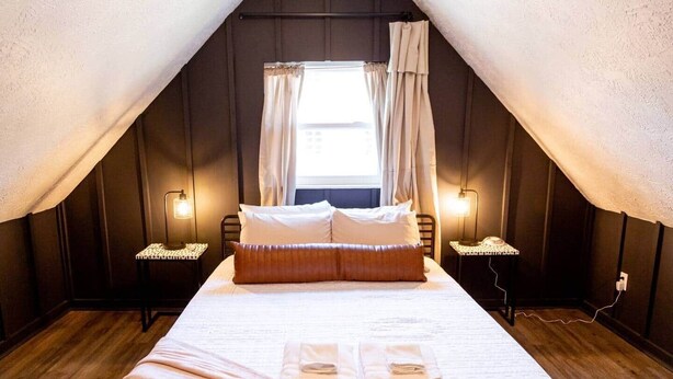 Une chambre confortable située sous les toits, avec un grand lit, deux tables de chevet, une fenêtre avec des rideaux et un plancher en bois.