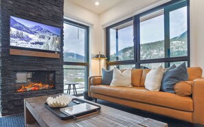 Una sala de estar con chimenea de piedra, sofá de cuero y ventanas amplias con vista a la montaña.