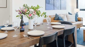 A dining area with a wooden table set for four, surrounded by black chairs. A vase with flowers is placed on the table, and there are wine glasses and plates ready for a meal.