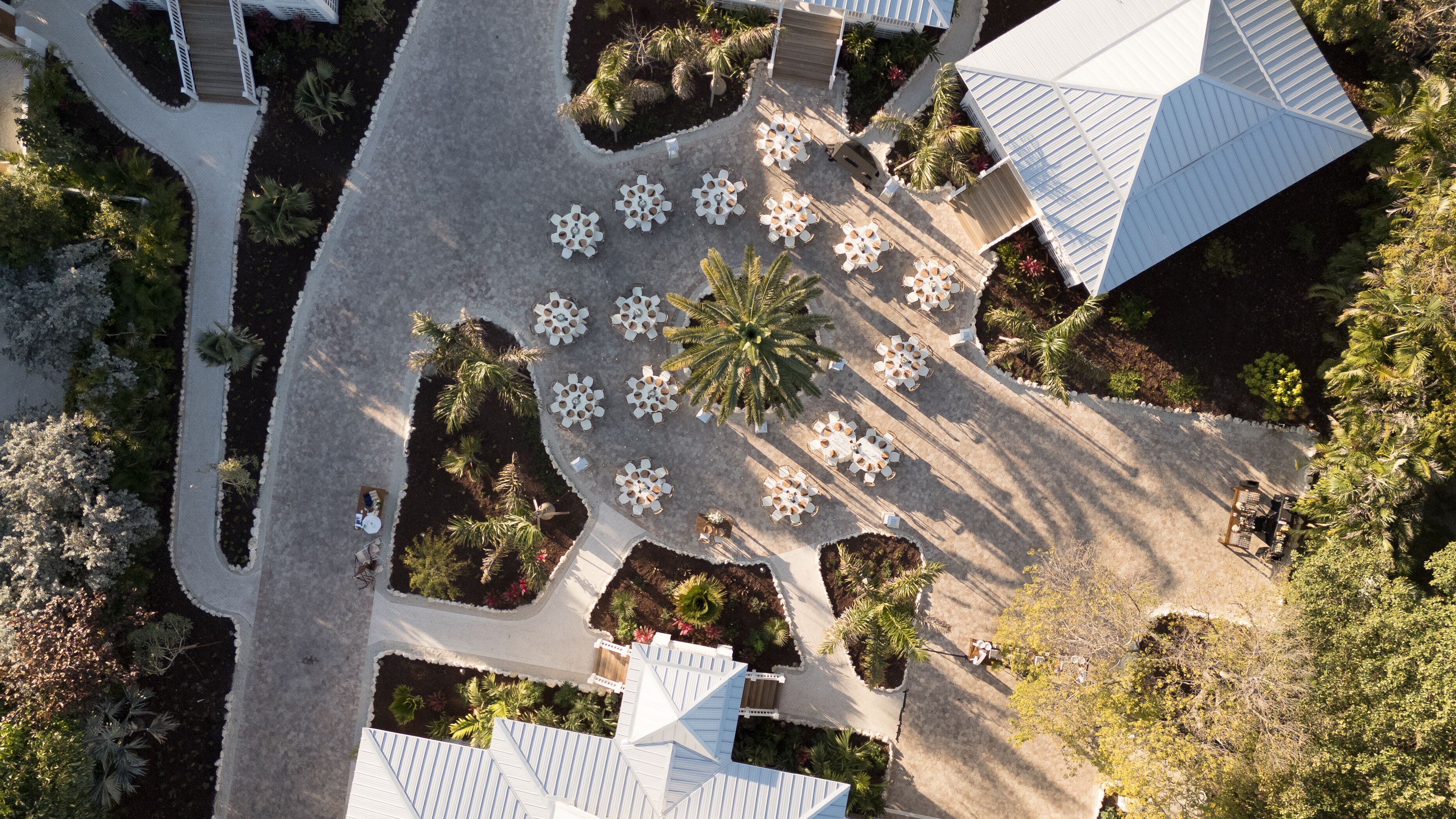 An aerial view of a resort with multiple white-roofed buildings, arranged in a circular pattern with outdoor seating areas and palm trees.