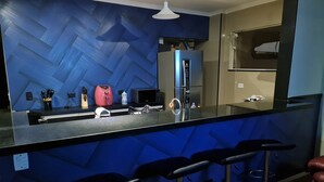 A modern kitchen with a blue herringbone backsplash, a stainless steel refrigerator, a microwave, and a sink. There are three black bar stools and a hanging light fixture above the counter. - Beachfront accommodation on Astúrias Beach. Spacious and comfortable. WiFi. (Guarujá)