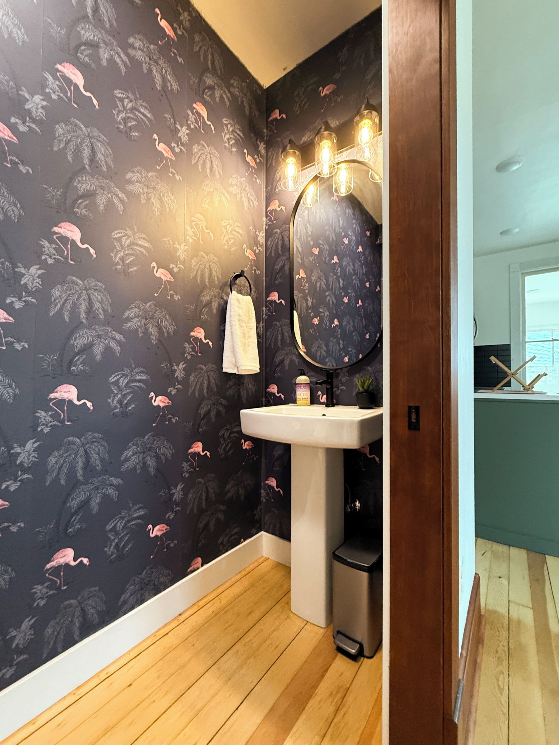 A bathroom with a dark floral wallpaper, a white sink, and a mirror with lights.
