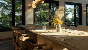 Uma sala de jantar com uma longa mesa de madeira posta para quatro pessoas, cercada por grandes janelas que dão vista para árvores.