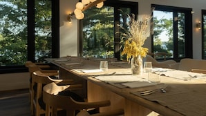A dining area with a long wooden table set for four, surrounded by large windows offering a view of trees. - Nourish + Reset at Dimensions | Cabin Ten | Breakfast Included (The Blue Mountains)
