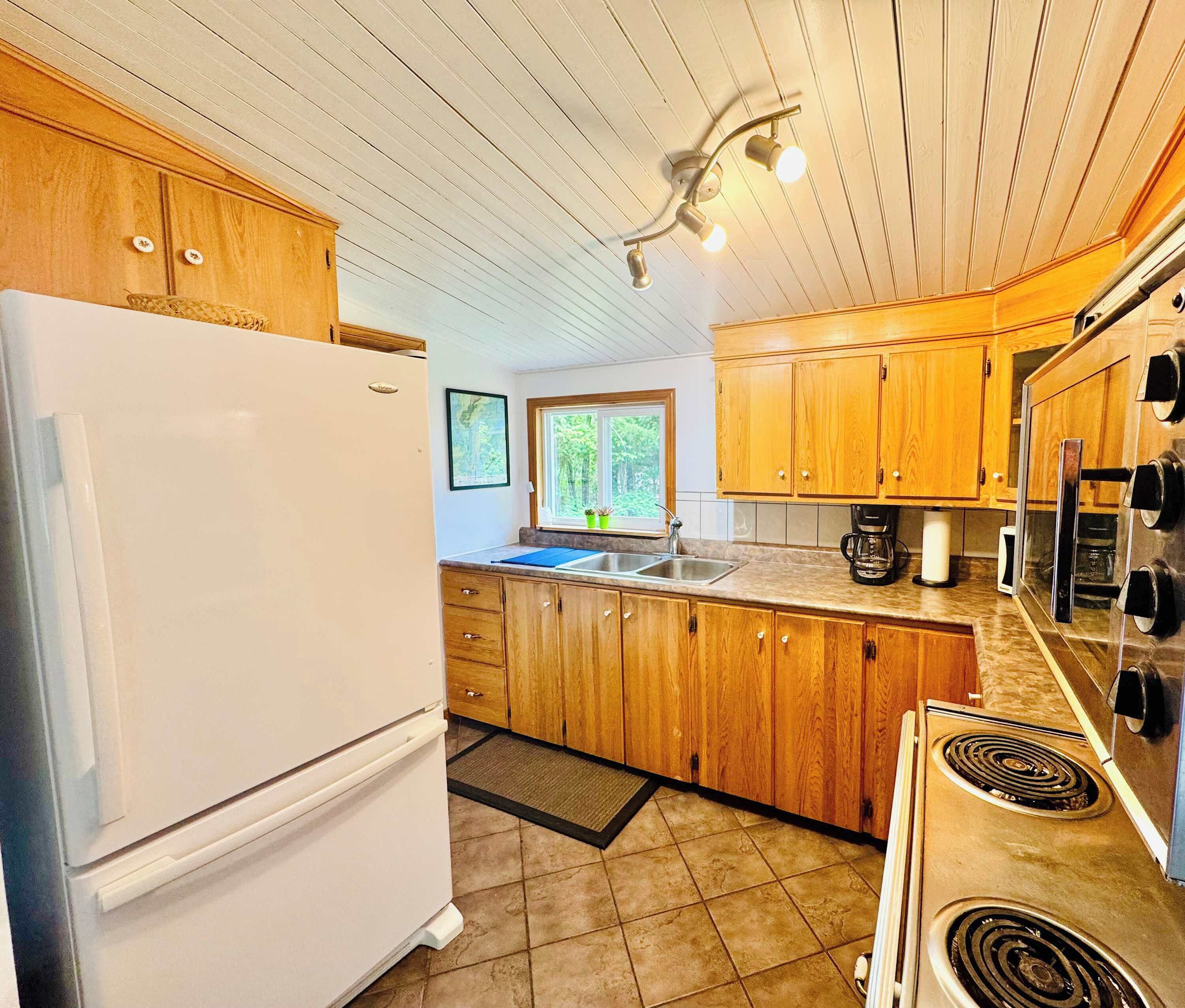 A kitchen with wooden cabinets, a white refrigerator, a stove, and a window with a view of trees.