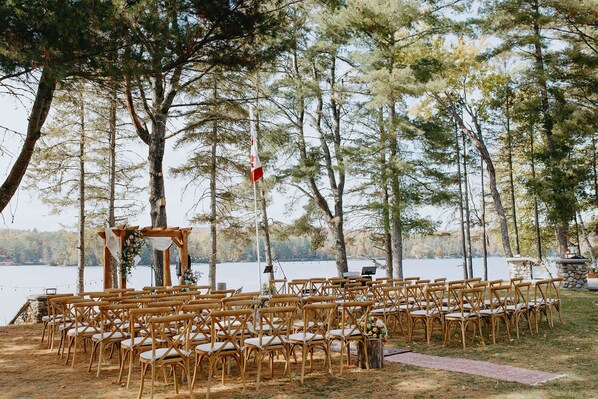An outdoor wedding ceremony setup with wooden chairs arranged in rows, a wooden arch, and a flagpole with a flag.