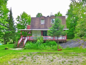 A two-story house with a porch and a small deck, surrounded by lush greenery and trees.