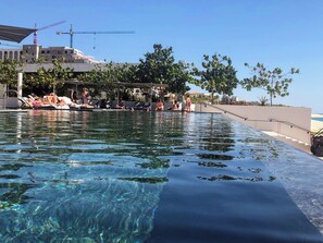 Una piscina con agua cristalina, rodeada de sillas de descanso y sombrillas, con vistas a edificios y árboles al fondo.