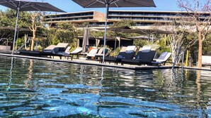 A pool area with lounge chairs and umbrellas, a clear pool, and a modern building in the background. - The Club at Solaz is a Luxury Villa with a beautiful view of the Sea of Cortez. (San José del Cabo)