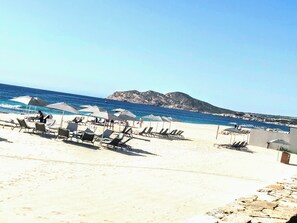 Una playa con tumbonas y sombrillas, cielo despejado y una montaña al fondo.