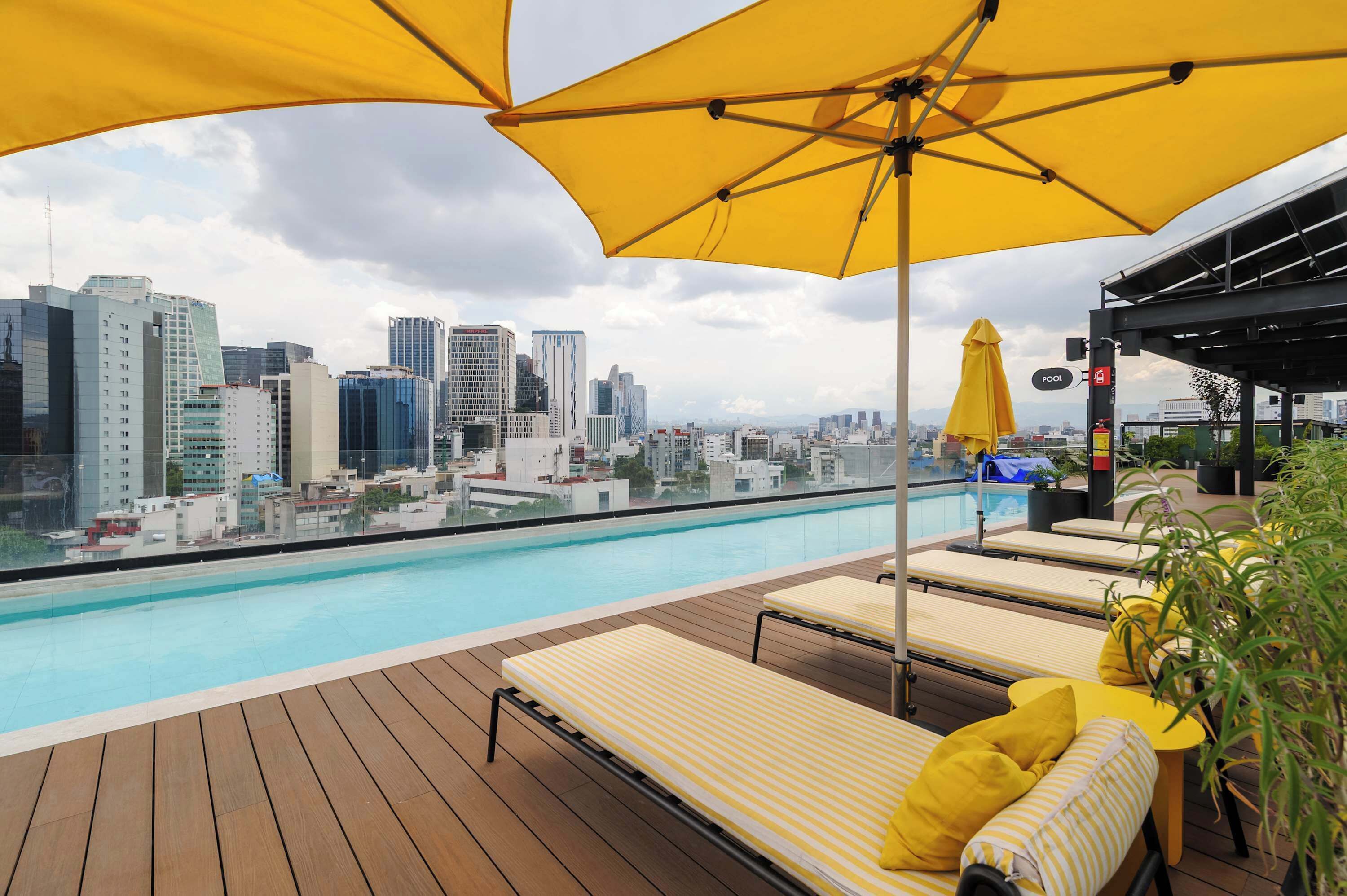 Un área de piscina en la azotea con sillones y sombrillas, con vistas a un paisaje urbano de edificios modernos.