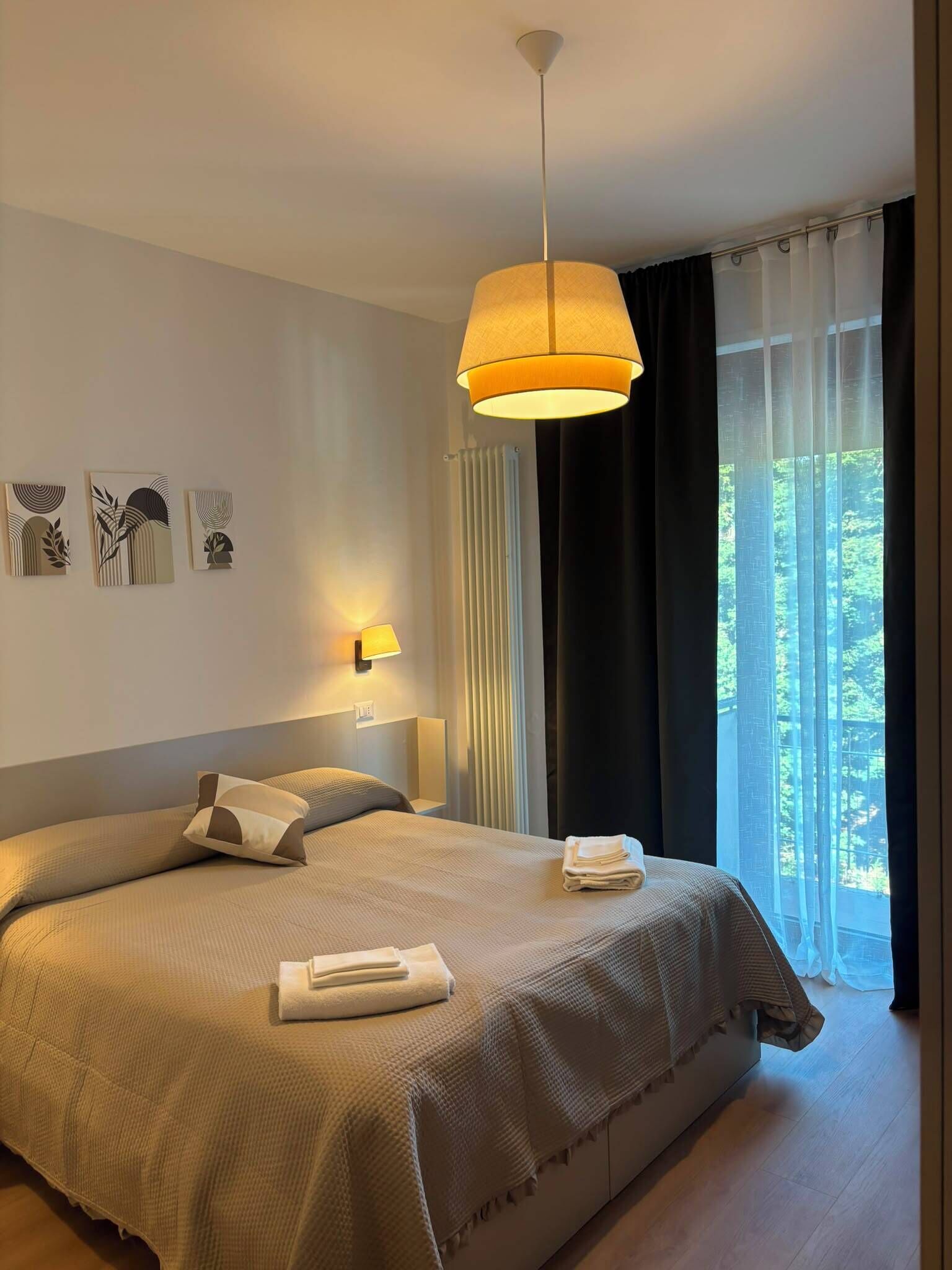 Une chambre d’hôtel avec un grand lit, des lampes de chevet, une vue sur la verdure à travers des rideaux transparents et des œuvres d’art abstraites accrochées aux murs.