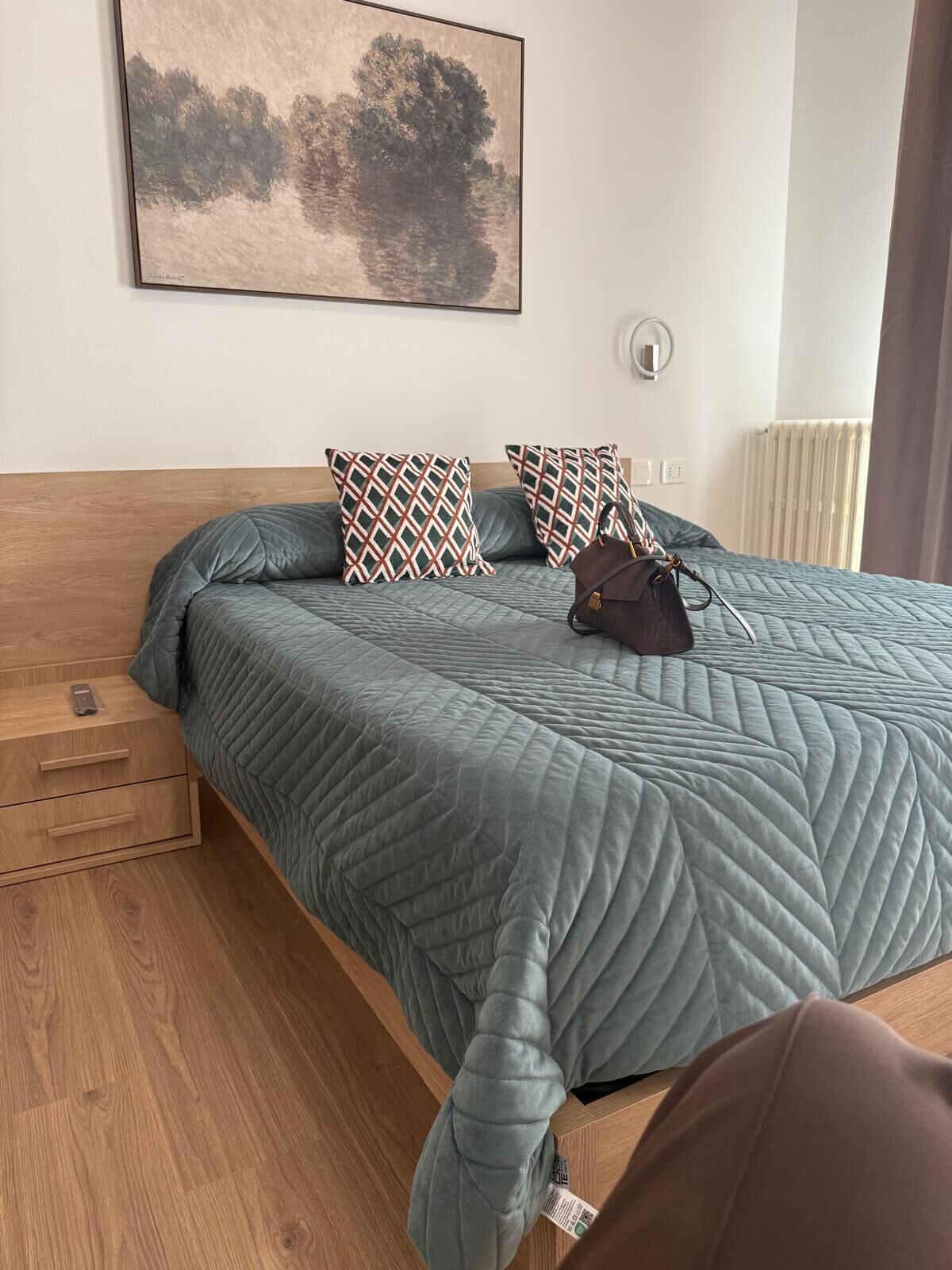 A neatly made bed with a quilted grey bedspread, two patterned pillows, a brown handbag, and a painting on the wall.