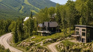 Une maison à deux étages, dotée d’un porche abrité et d’une extension moderne, entourée d’un paysage boisé et bordée d’un chemin de terre sinueux.