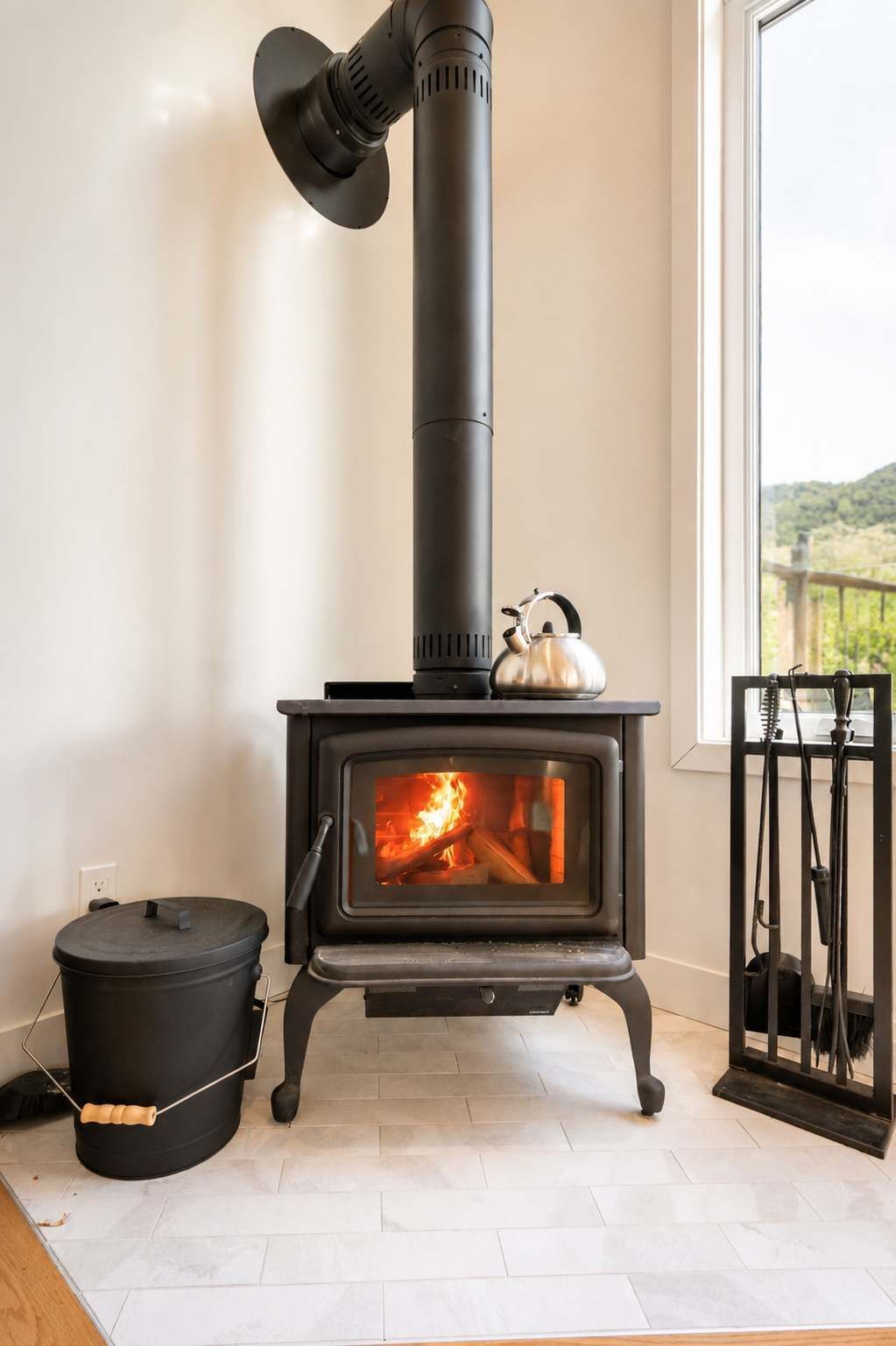 Un poêle à bois avec une cheminée, une bouilloire posée dessus, et une vue sur l’extérieur par une fenêtre.