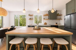 Una cocina moderna con una isla grande, taburetes de bar, iluminación colgante y vistas al exterior.