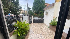 Un patio piastrellato con un cancello in metallo, un piccolo giardino e un muro bianco con un armadietto tecnico.