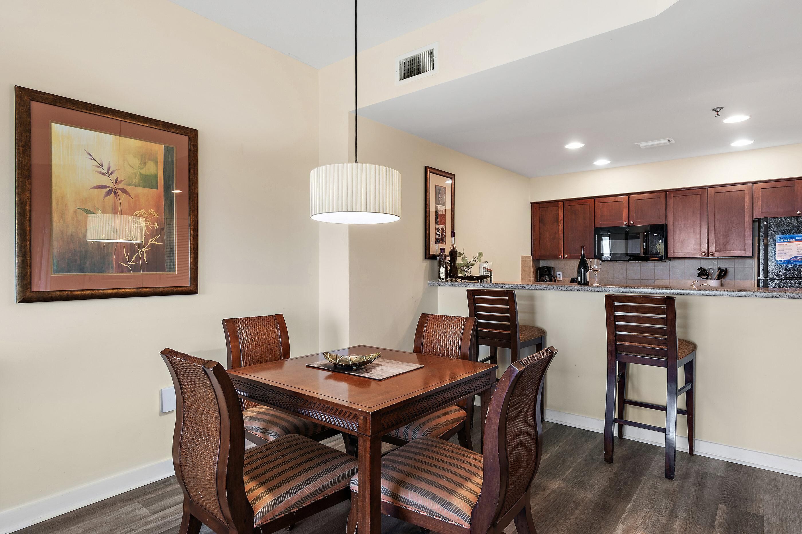 A dining area with a wooden table and chairs, a hanging light fixture, and a kitchen in the background.