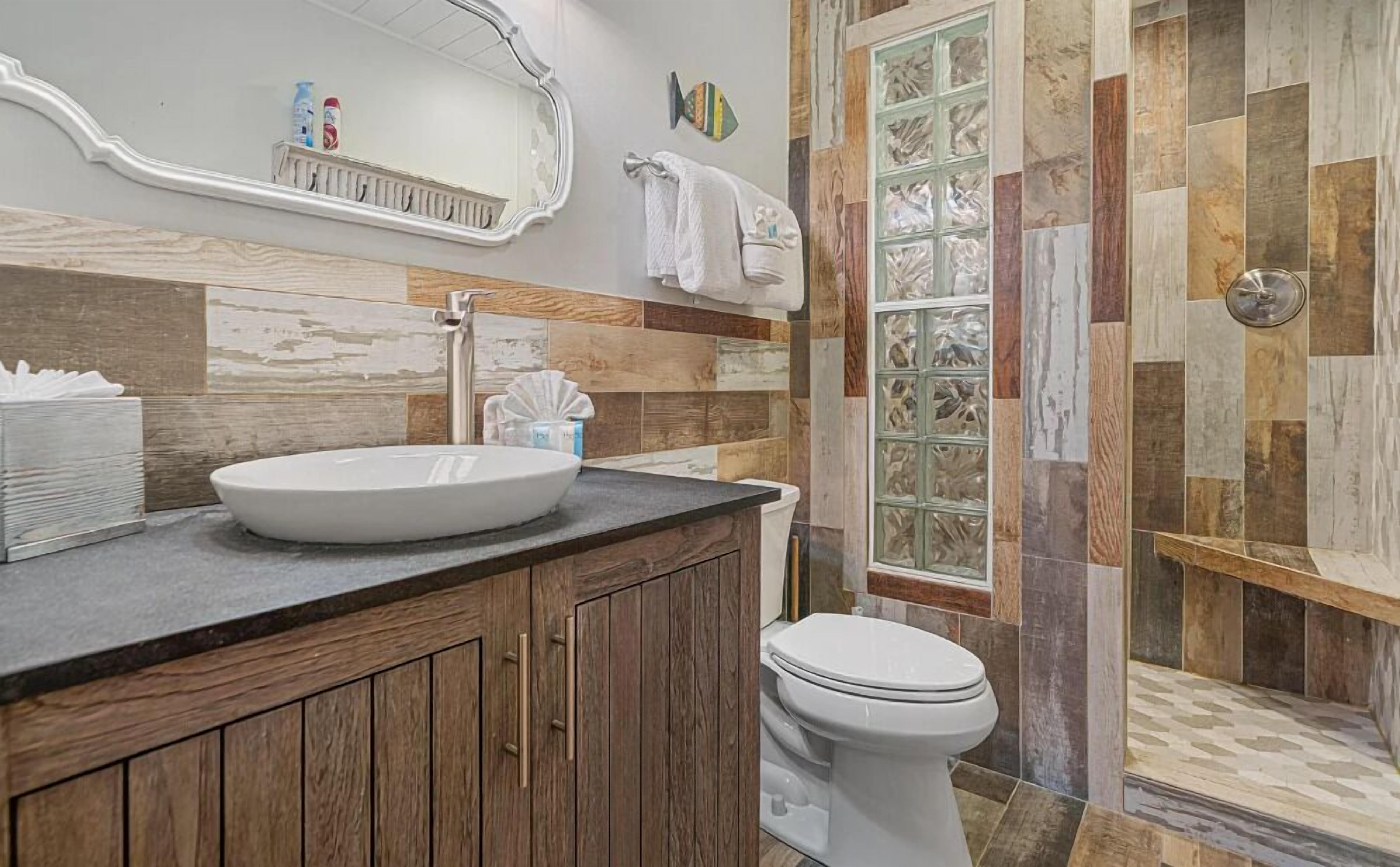 Un baño con tocador de madera, lavamanos blanco, inodoro y ducha con puerta de vidrio.
