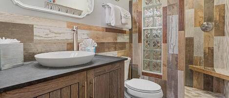 Un baño con tocador de madera, lavamanos blanco, inodoro y ducha con puerta de vidrio.