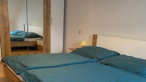 Een houten bed met blauwgroene beddengoed, een houten kledingkast en een nachtkastje met een lamp.