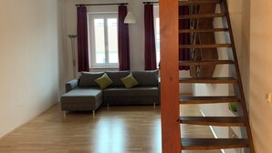 Een ruime woonkamer met een houten trap, een grijze bank met groene kussens, en een groot raam met gordijnen.