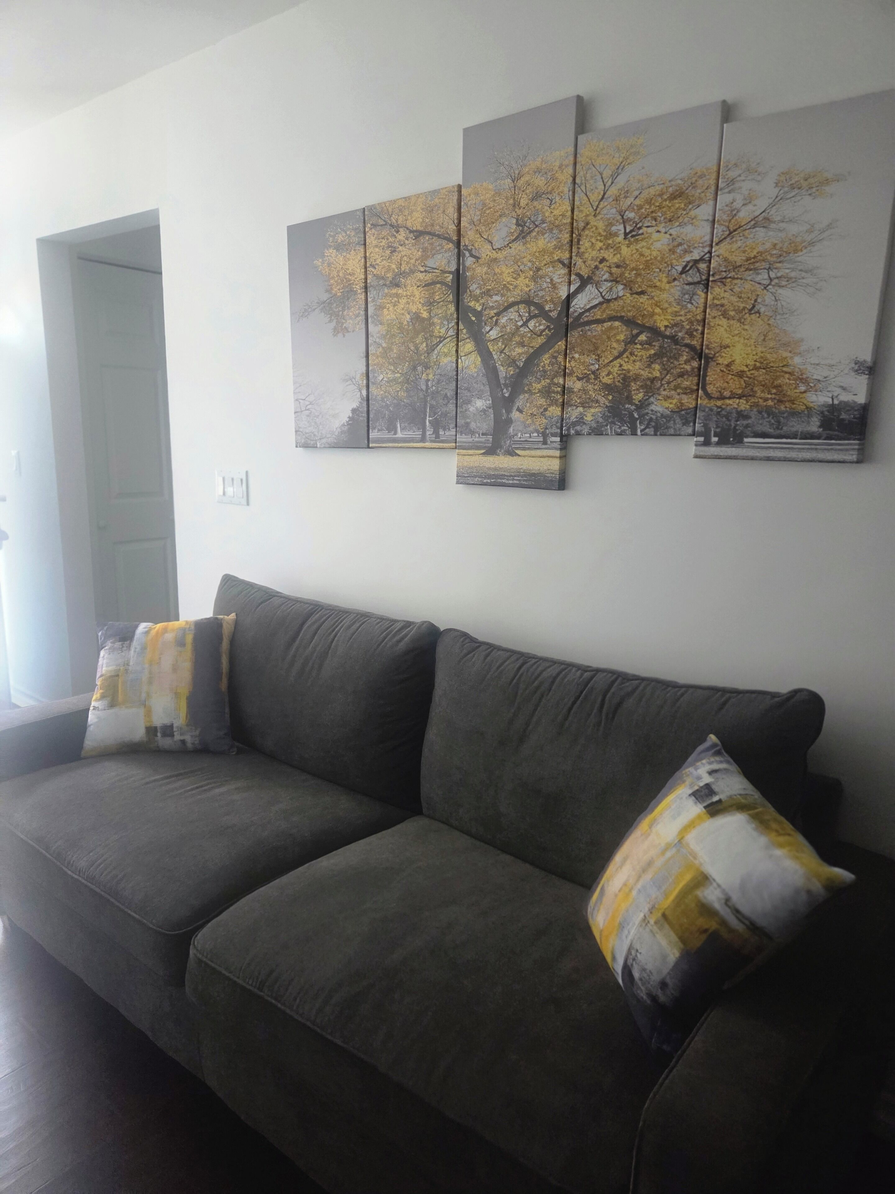 A living room with a gray sofa, two cushions, and a wall-mounted triptych painting of a tree.
