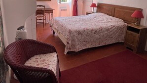 Un dormitorio con cama, una silla, un escritorio, una ventana con cortinas, un cuadro y una lámpara.