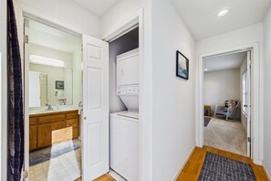 A hallway with a white door leading to a bathroom with a sink and mirror, a laundry area with a washing machine, and a chair in the corner.