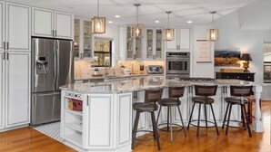 A modern kitchen with white cabinetry, stainless steel appliances, a large island with bar stools, pendant lighting, and a view of the outdoors. - Peaceful Riverfront Retreat with Stunning Views (Mountain Home)