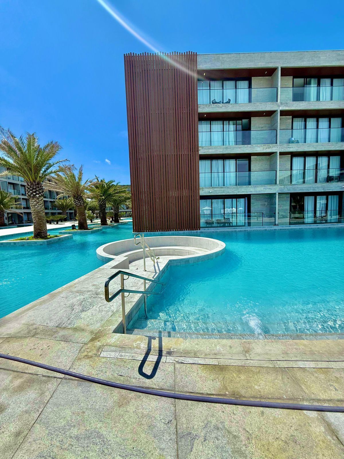A modern hotel with a pool area, featuring a large rectangular structure in the foreground, a multi-story building in the background, and palm trees around the pool.