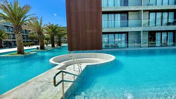 Un hotel moderno con zona de piscina, destacando una estructura rectangular grande al frente, un edificio de varios pisos al fondo y palmeras alrededor de la piscina.