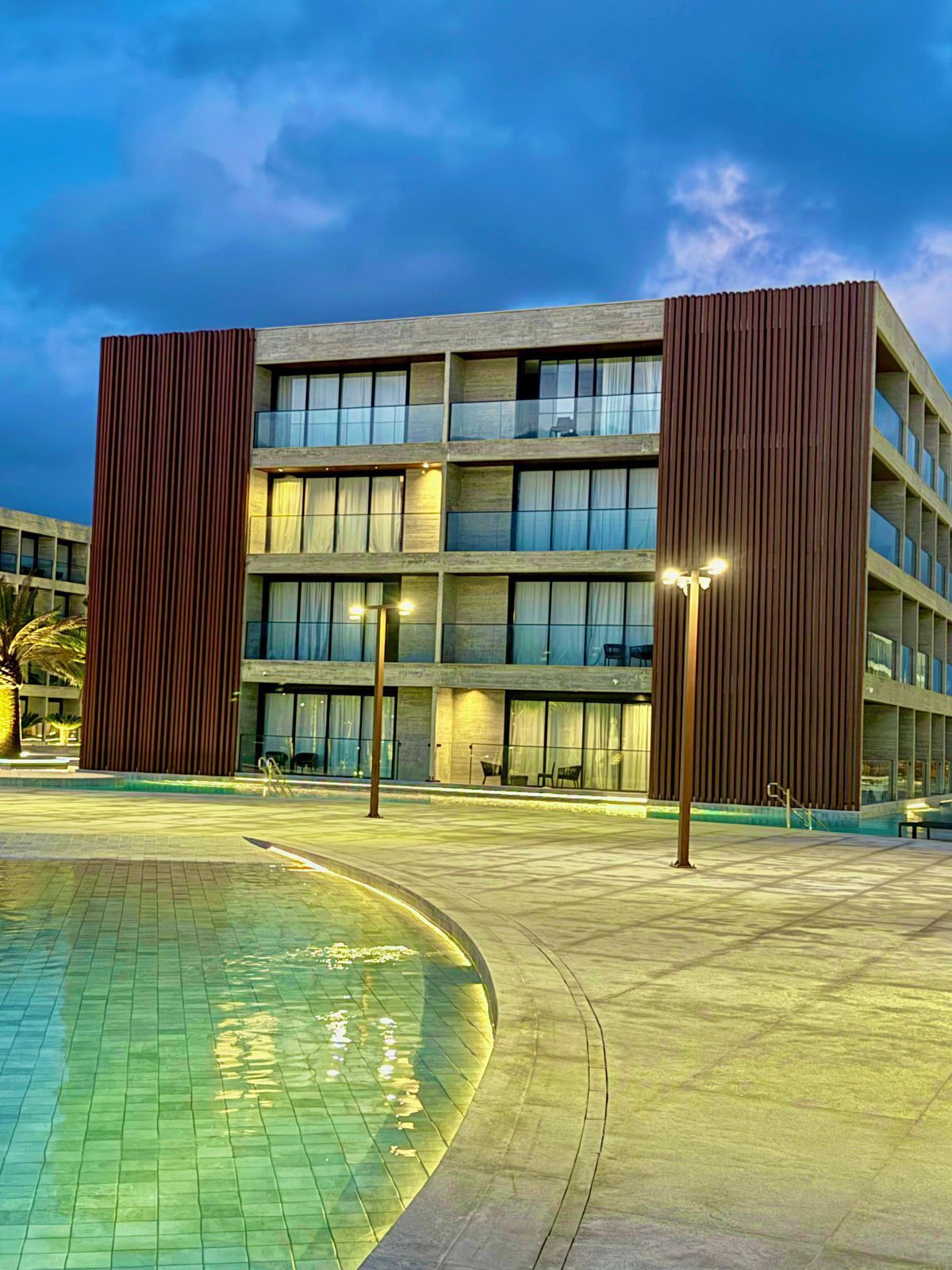 Un bâtiment moderne doté d’un espace piscine comprenant une grande piscine, un espace salon couvert et une entrée bien éclairée.