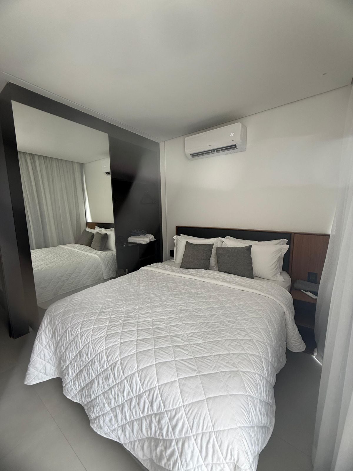 Una habitación de hotel moderna con una cama grande, una mesita de noche, un espejo y un equipo de aire acondicionado en la pared.