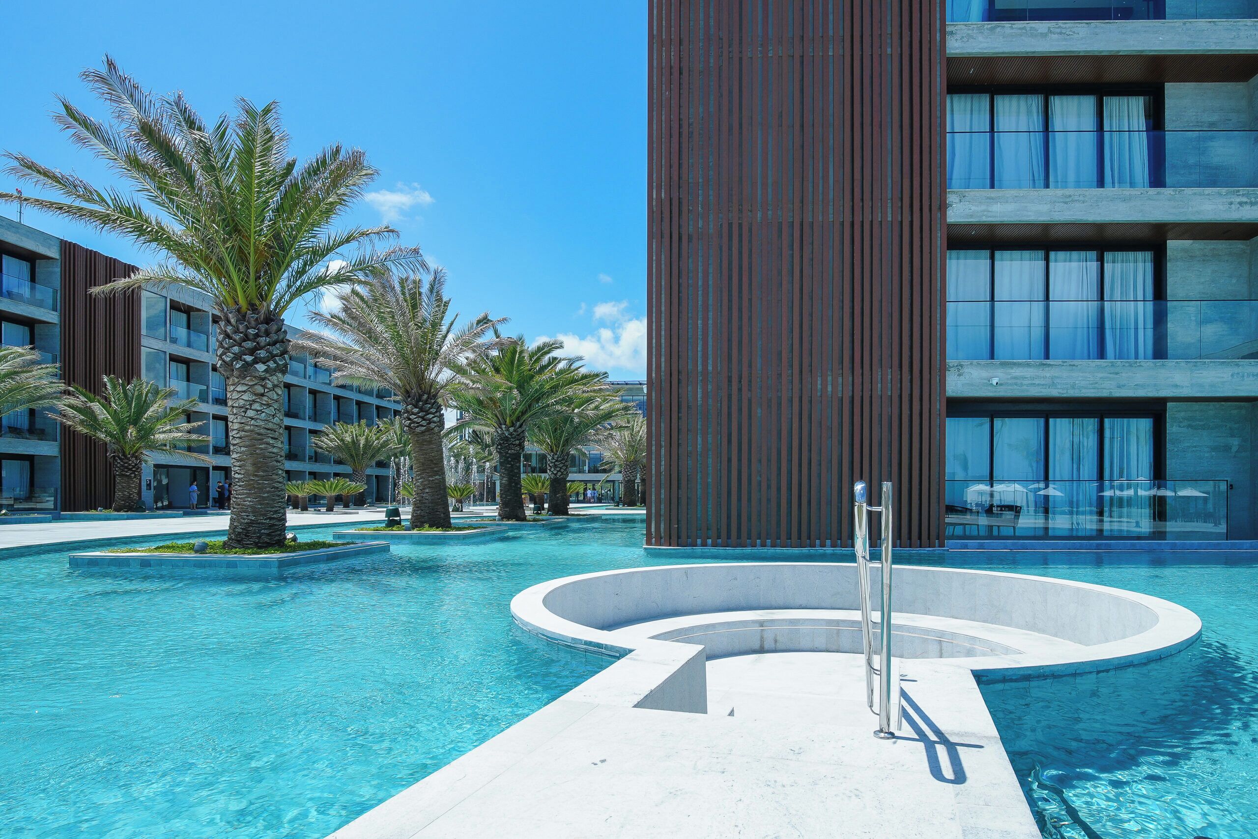 Un hotel moderno con piscina, palmeras y un edificio alto con paneles de madera verticales.