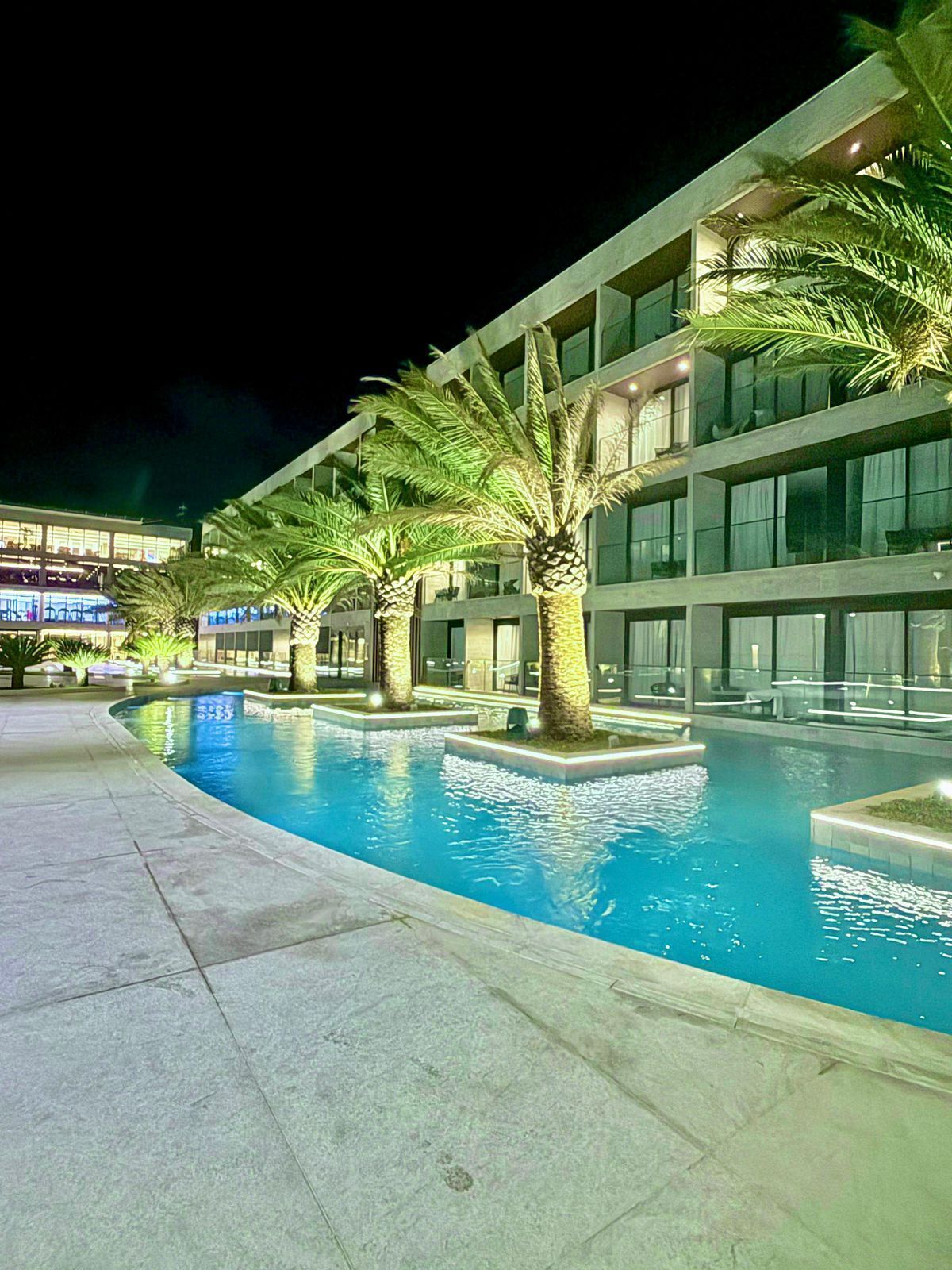 A nighttime swimming pool area with palm trees and a modern building with multiple floors and large windows.