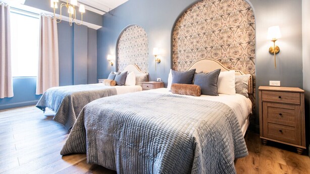 A bedroom with two beds, a chandelier, patterned wallpaper, and a nightstand with a lamp. - Hollyhock Suite 406: 1 bed/1 bath (Louisville)
