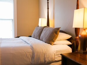 A neatly made bed with white linens and two pillows, a wooden nightstand with a lamp, and a window with blinds. - Deluxe Room 7-Double room-Private Bathroom-Standard (Fish Creek)