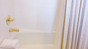A white tiled bathroom with a bathtub, a gold faucet, and a towel stack.