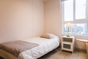 A minimalistic bedroom with a bed, a nightstand, a lamp, and a view of a cityscape through a window. - Beautiful Apartment in Nunoa with Balcony and Pool (Ñuñoa)