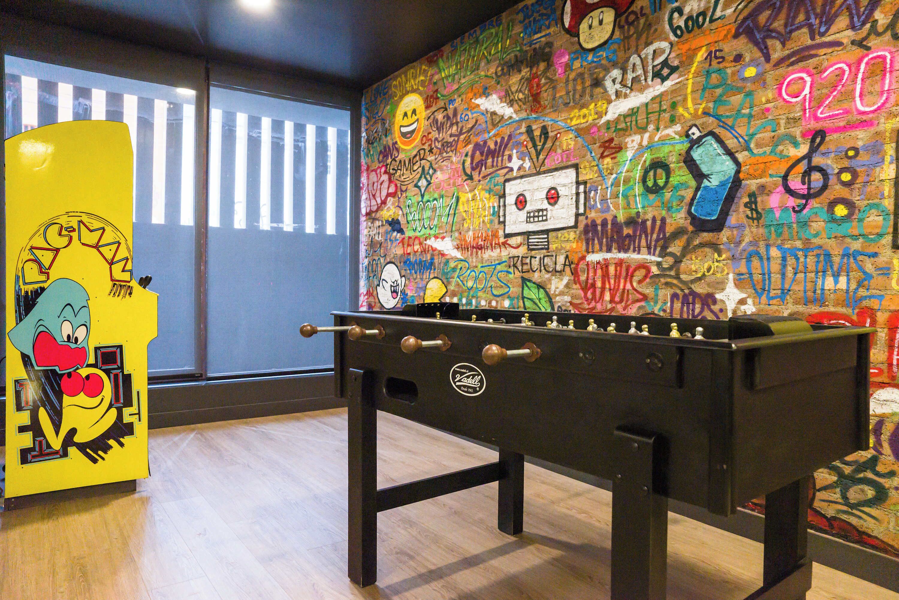 Une table de baby-foot dans une pièce dont un mur est couvert de graffitis et qui possède une fenêtre avec des stores.