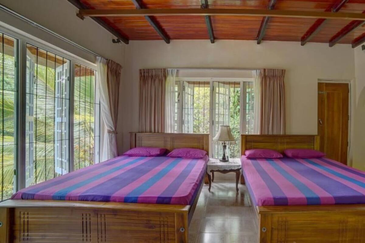 A room with two beds, a wooden ceiling, and large windows with curtains.