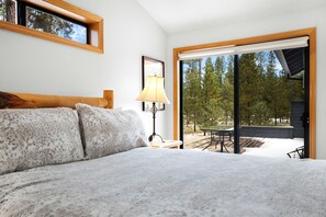 A bedroom with a large bed, a nightstand, a lamp, and a view of the outdoors. - Sunriver's Klamath Lane - Hot Tub & SHARC Passes (Sunriver)