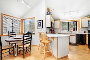 A modern kitchen with a dining area, wooden chairs, a white island, and a large window with blinds. - Sunriver's Klamath Lane - Hot Tub & SHARC Passes (Sunriver)
